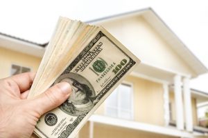 Hand holding money with a house in the background.