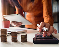 Woman using calculator, with notebook, laptop, and stacks of coins.