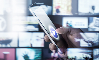 Hand holding a cell phone with a wall full to televisions in the background.