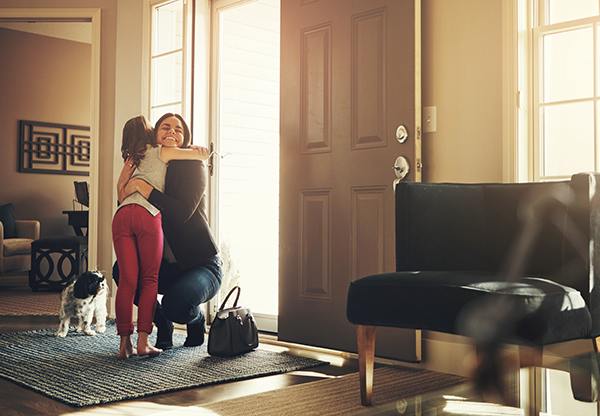 Mother coming home from work hugging a young child inside by the open front door.