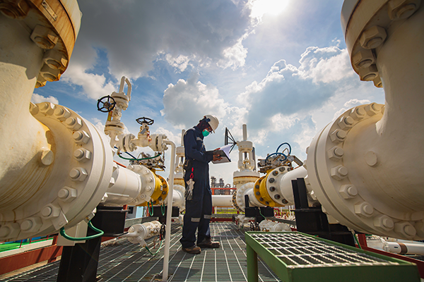 Oil and gas worker in uniform looking at a clipboard surrounded by pipelines.