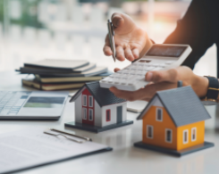Person holding calculator, with models of houses and clipboard.