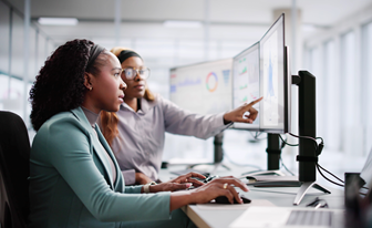 Two female analysts looking at market data on multiple computer screens.