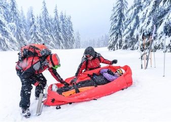 Two ski patrollers with a rescue sled helping an injured woman on a snowy trail.
