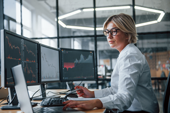 Female stock broker analyzing financial data on multiple computer screens. 