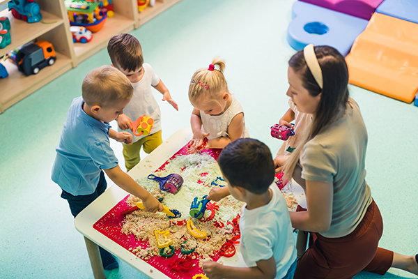 Childcare worker and five children around a table playing with kinetic sand