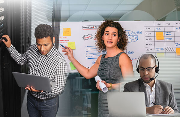 Two men use laptops. One of these men wears headphones. A woman holds a rolled-up paper in one hand and a pointer in the other. She uses the pointer to point to a section of a whiteboard. The whiteboard has a figure, a table, and other writing. Some sticky notes are stuck to the whiteboard.